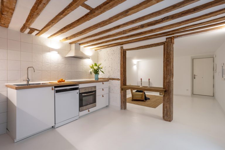 Vista de amplia cocina con vigas de madera vistas. Muebles blanco con tiradores de cuero. mandarinas y jarrón con flores sobre la encimera.