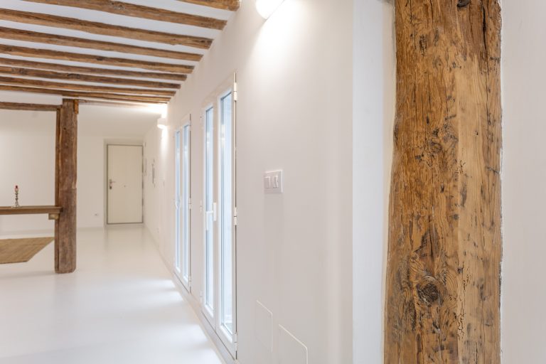 Vista de pasillo con vigas de madera y techo con vigas vistas. Ventanas a patio y puerta de entrada de vivienda.