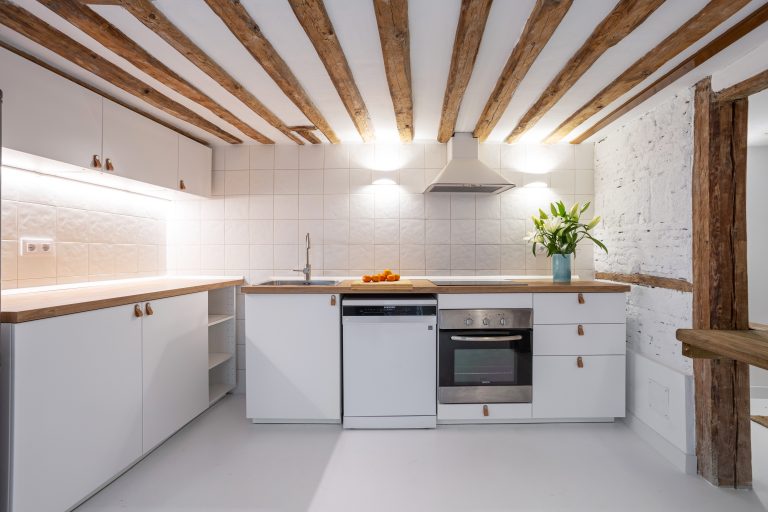 Vista general de cocina con muebles blancos y techo con vigas de madera vistas. Mandarinas sobre la encimera y jarrón con flores blancas.