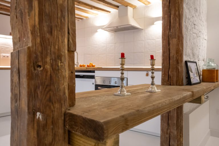 Vista de muebles de cocina, con jarrón con flores blancas. Vigas con mesa rústica y candelabros con vela rojas, jarrones y cuadro apoyado sobre la mesa.