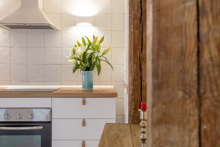 Vista de lateral de muebles de cocina, con jarrón con flores blancas. Vigas con mesa rústica y candelabro con vela roja.