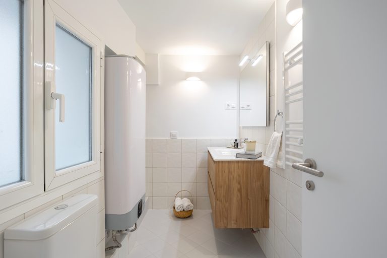 Vista completa de baño con inodoro, termo eléctrico, lavabo acabado roble y radiador toallero. Puerta de paso abatible color blanco.