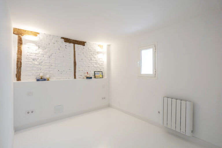 Cabecero de cama de obra con ladrillos vistos pintados de blanco y vigas vistas en madera. Libros, velas y un cuadro con una lámina en acuarela.