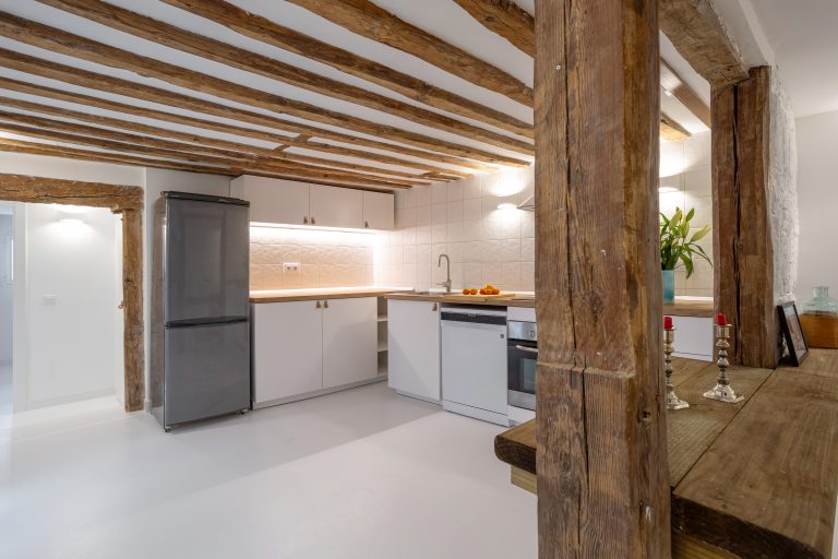 Vista de amplia cocina con vigas de madera vistas. Muebles blanco con tiradores de cuero. mandarinas y jarrón con flores sobre la encimera.