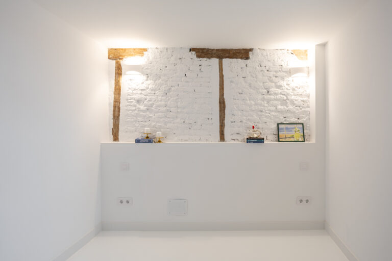 Cabecero de cama de obra con ladrillos vistos pintados de blanco y vigas vistas en madera. Libros, velas y un cuadro con una lámina en acuarela.