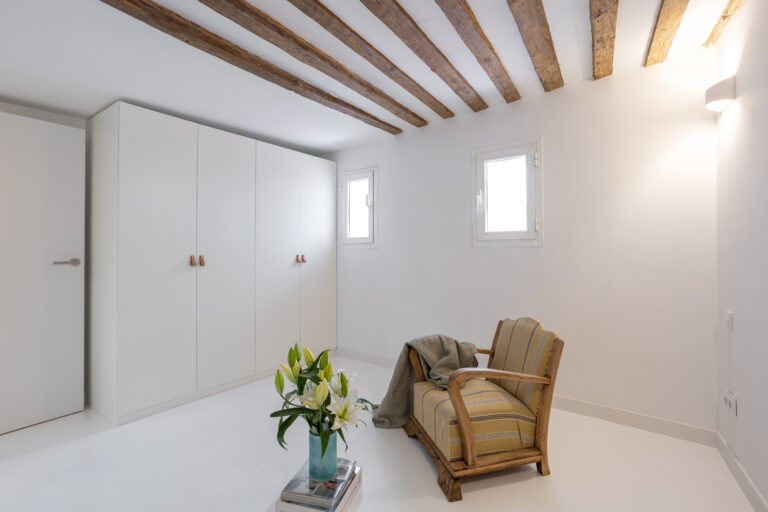 Armario blanco de Ikea en habitación blanca con vigas de madera, flores blancas y sillón estilo rústico.
