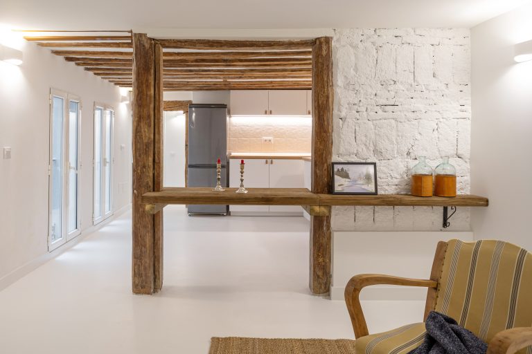 Mesa de madera rústica apoyada entre vigas. Muro con ladrillos vistos pintados en color blanco. Salón luminoso y muebles de cocina en color blanco y encimera de madera.