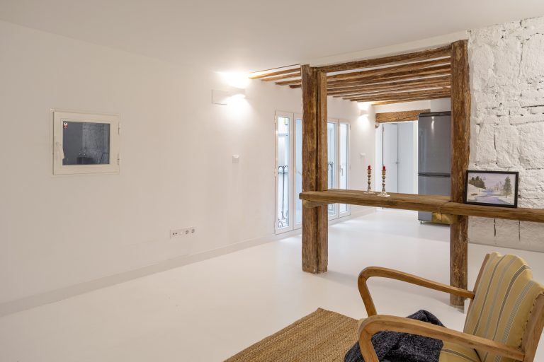 Mesa de madera rústica apoyada entre vigas. Muro con ladrillos vistos pintados en color blanco. Salón luminoso y muebles de cocina en color blanco y encimera de madera.