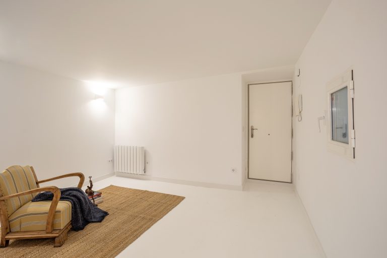 Amplio salón con paredes y suelo blanco. Sillón estilo rústico y alfombra de fibra natural