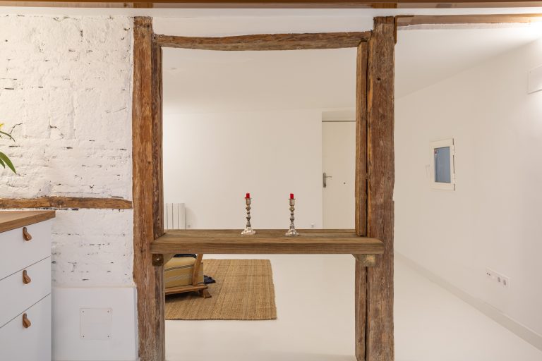 Mesa de madera rústica apoyada entre vigas. Muro con ladrillos vistos pintados en color blanco. Salón luminoso y muebles de cocina en color blanco y encimera de madera.